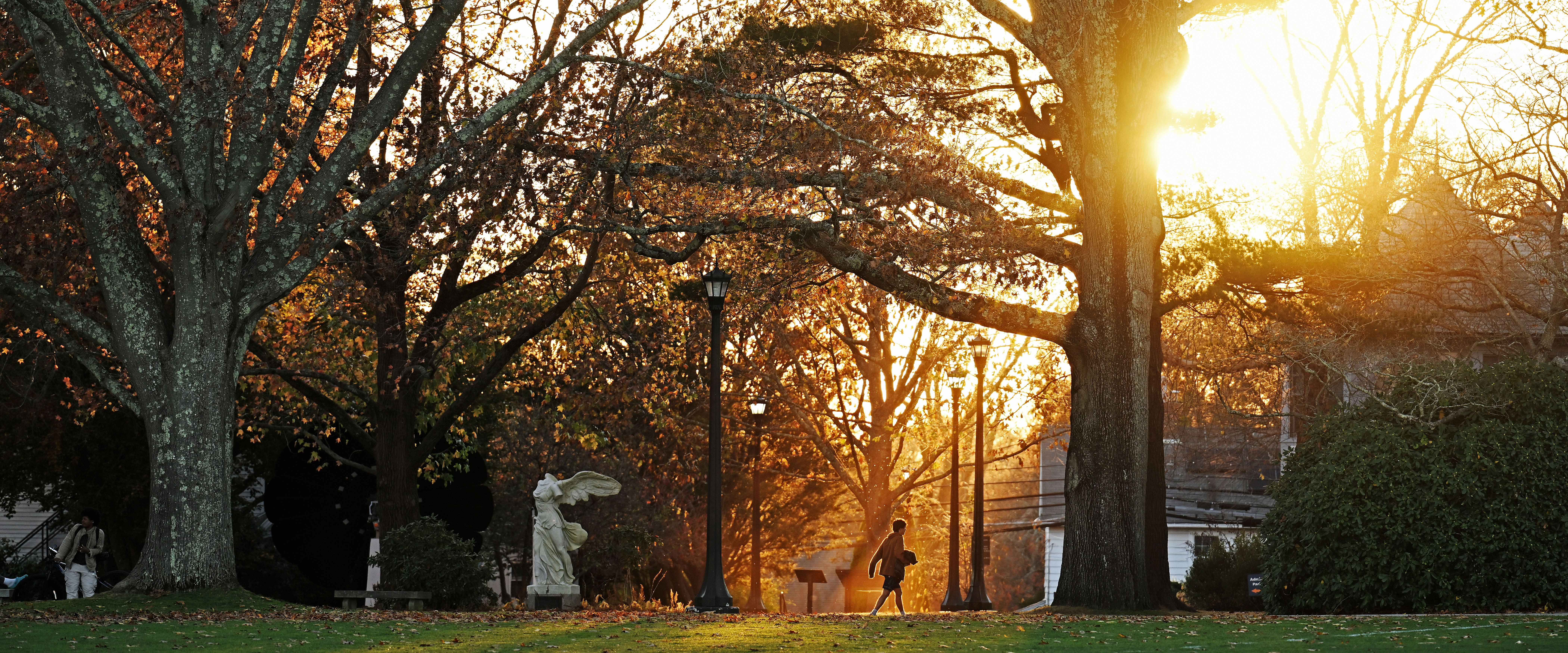 A lone students walks across campus as the sun sets behind the trees.