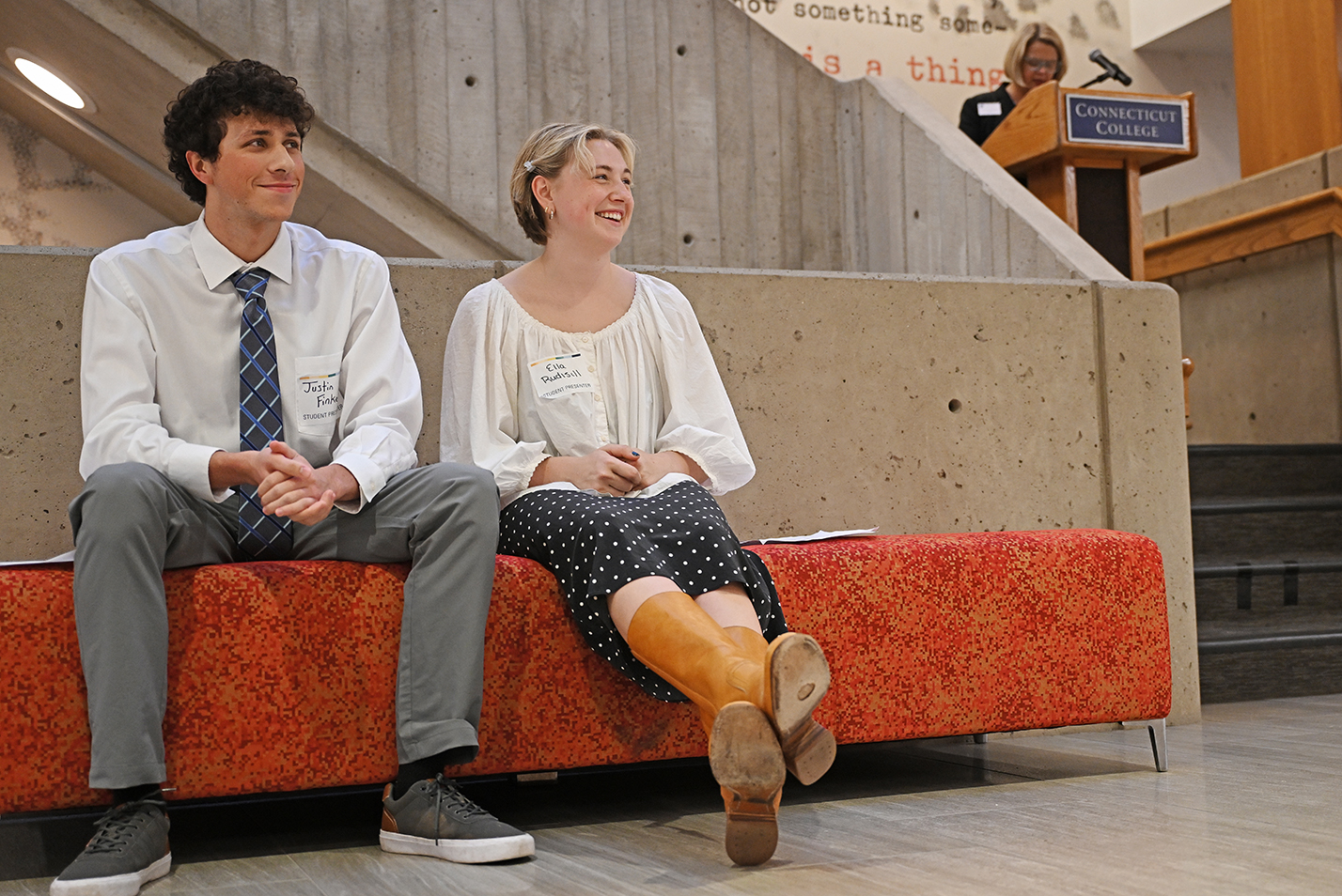 Two well-dressed students sit on a cushioned bench waiting for their turn at the podium while the college president makes remarks.