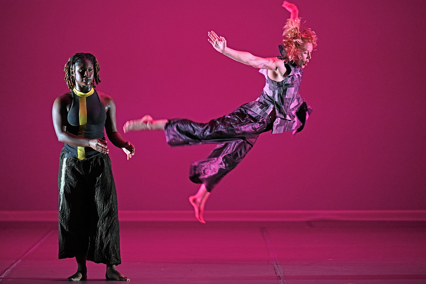 One dancer leaps into the air as another strikes a static pose to the side during a modern dance performance.