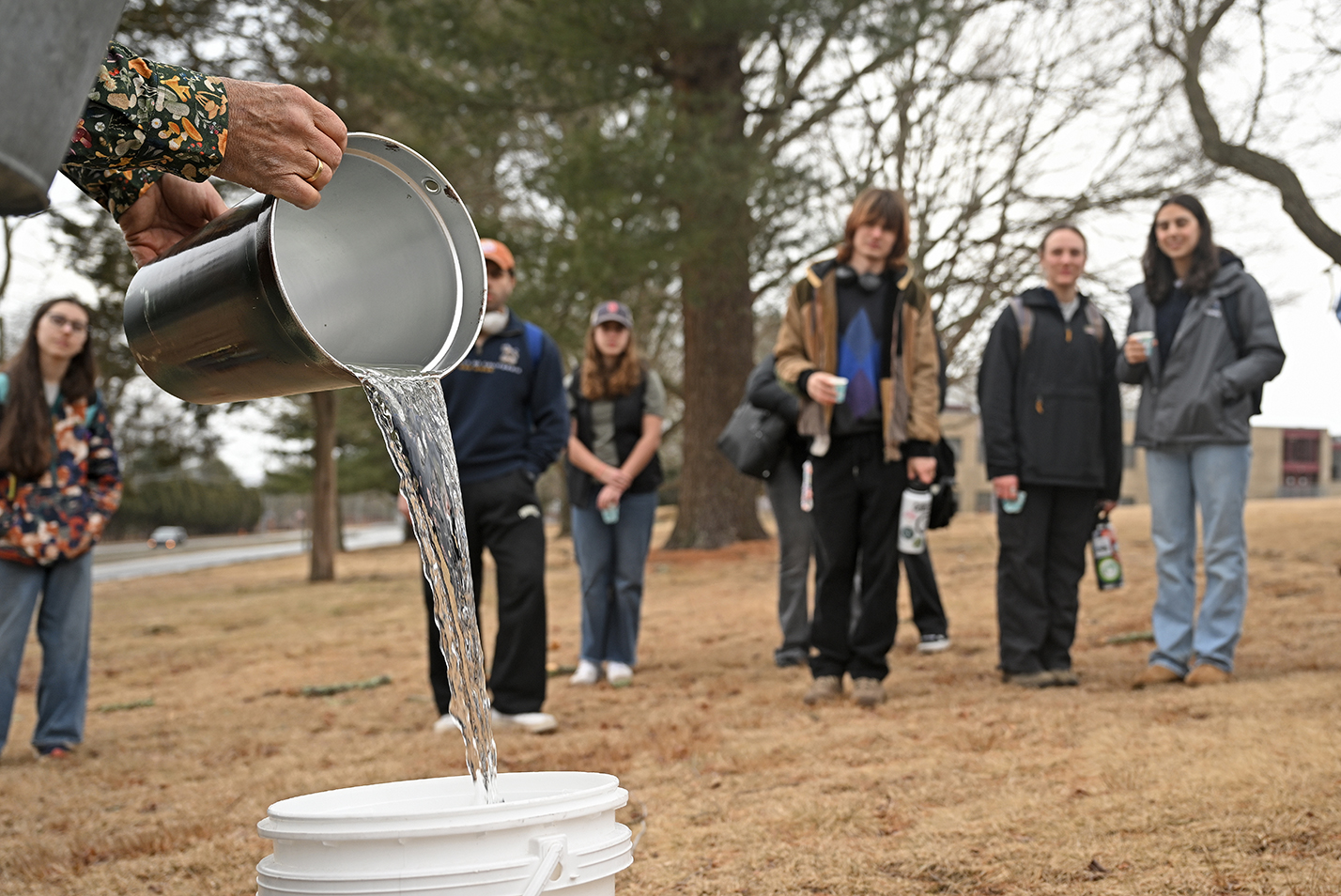 A college professor pours sap collected from a maple tree from the collection bucket as students watch.