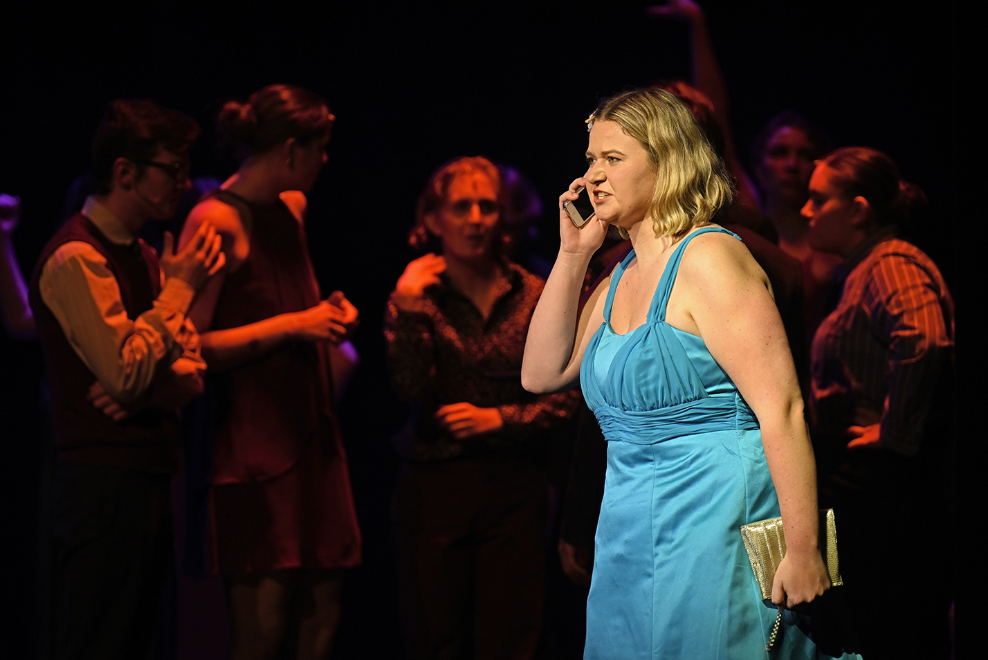 a female actor in a blue dress speaks angrily into a cell phone prop during a musical theater performance.