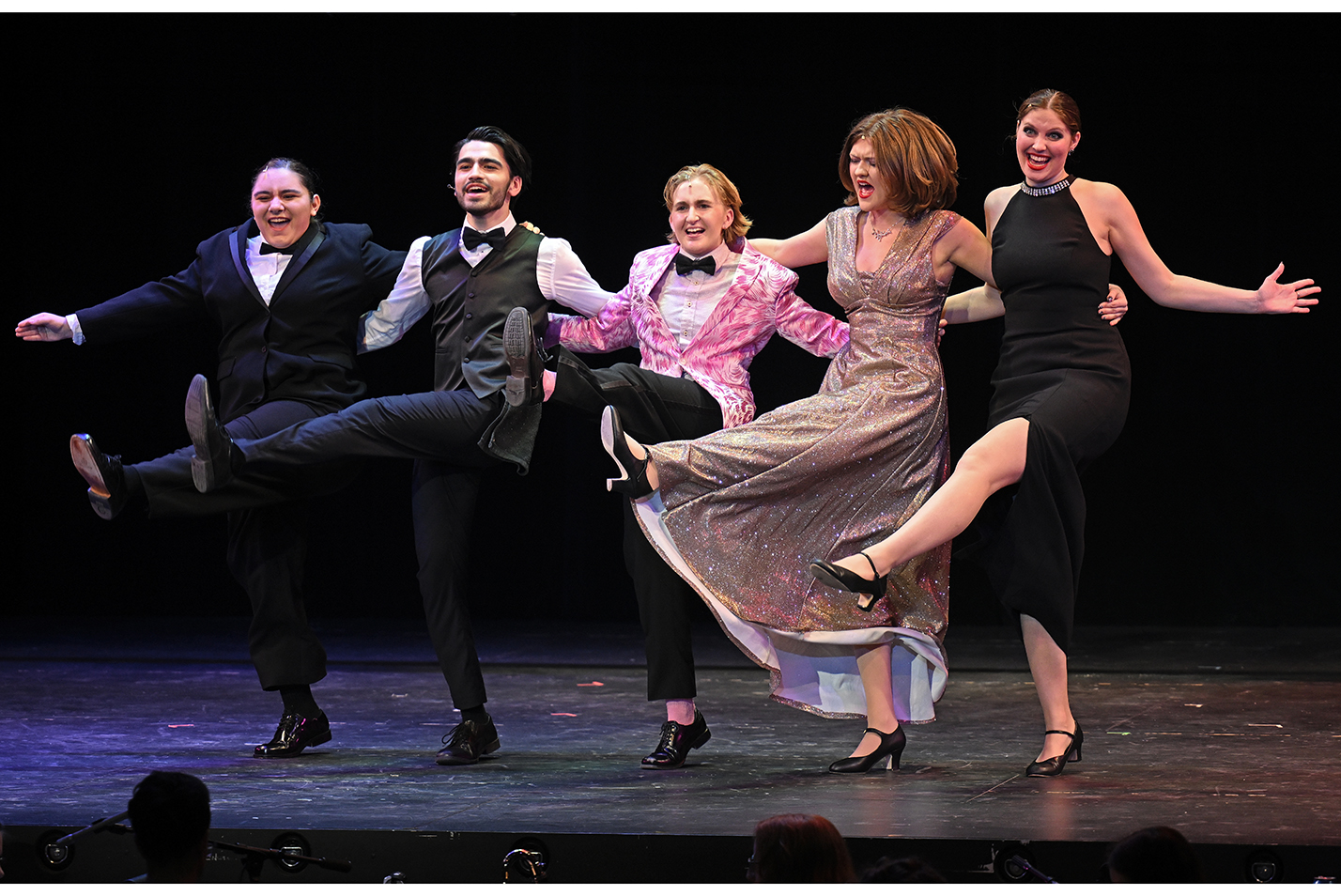 five actors in fancy costumes perform a kick line as part of a musical theater performance.